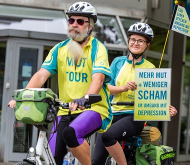 Zwei Personen sitzen auf einem Tandemfahrrad. Die hintere Person hält ein Schild mit der Aufschrift "Mehr Mut + weniger Scham im Umgang mit Depression"