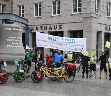 Mehrere Menschen stehen mit Fahrrädern vor einem Rathaus. Sie halten ein Banner und Schilder. Zwei Personen verdecken ihr Gesicht mit Smileys.
