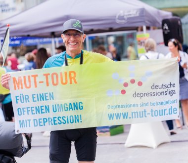 Eine Person hält ein Banner in die Kamera. Auf dem Banner steht „MUT-Tour Für einen offenen Umgang mit Depression!“
