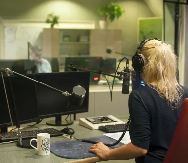 Eine Person mit langen Haaren sitzt in einem Radiostudio. Sie sitzt an einem Schreibtisch und trägt Kopfhörer.