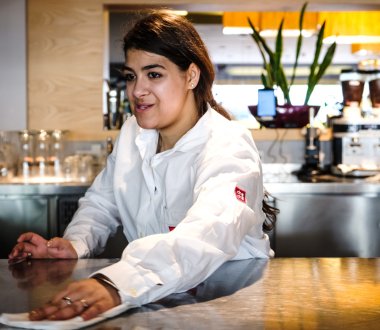 Eine Frau steht in Arbeitskleidung in einem Café und wischt den Tresen mit einem Tuch.