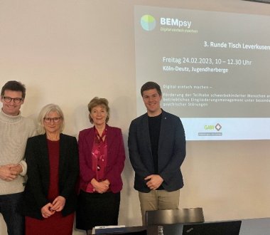 Vier Personen stehen vor einer Wand, an der eine Präsentation zum Projekt BEMpsy zu sehen ist.