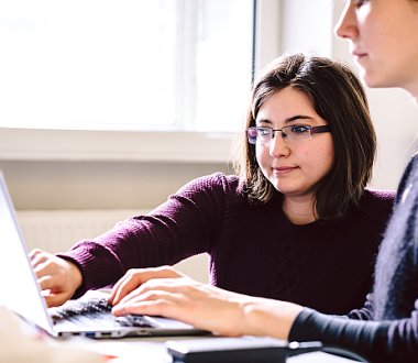 Zwei Personen sitzen an einem Tisch. Eine Person tippt, die andere Person zeigt auf den Laptopbildschirm.
