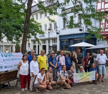 Viele Menschen stehen mit zwei bepackten Pferden auf einem Platz. Sie halten ein Banner hoch, auf dem MUT-Tour steht.