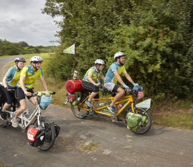 Auf einer nicht asphaltierten Strecke fahren zwei Fahrradtandems mit jeweils zwei Personen und vielen Taschen und Fähnchen.