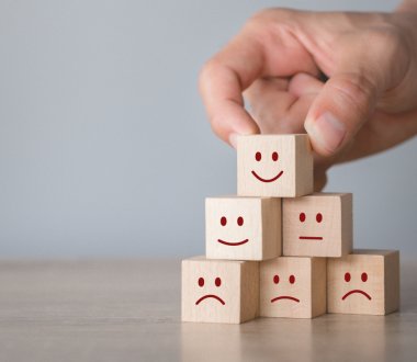 Eine Hand stapelt mehrere kleine Holzklötze zu einer Pyramide. Auf den Holzklötzen sind unterschiedliche Emotionen durch smileys dargestellt. Von glücklich bis traurig.