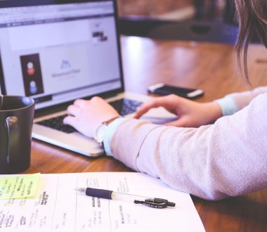 Eine Person sitzt an einem Laptop und tippt etwas. Neben ihr liegt auf dem Tisch ein beschriebener Schreibblock mit Stift.