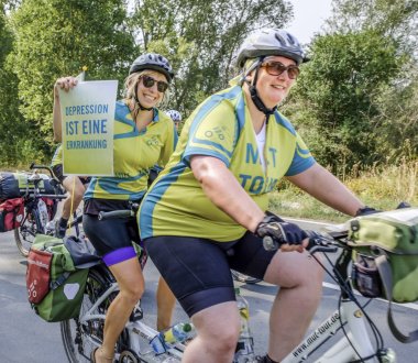 Zwei Personen fahren gemeinsam mit einem Tandem. Die hintere Person hält ein Schild hoch. Darauf steht "Depression ist eine Erkrankung".