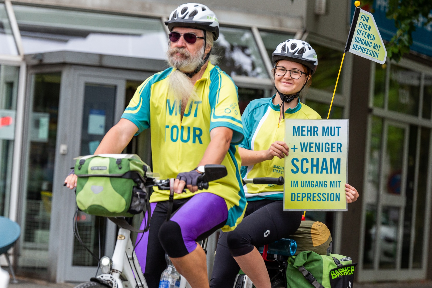 Zwei Personen sitzen auf einem Tandemfahrrad. Die hintere Person hält ein Schild mit der Aufschrift "Mehr Mut + weniger Scham im Umgang mit Depression"