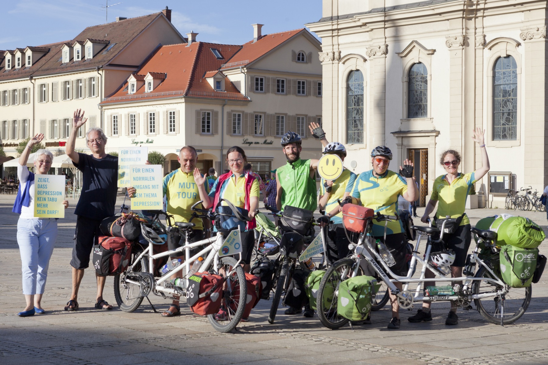 Mehrere Teilnehmer der MUT Tour mit Ihren Fahrrädern. Sie winken und halten Schilder mit Aufschriften wie "Zeigt, dass Depression kein TAbu mehr sein darf!" oder "Für einen normalen Umgang mit dem Thema Depression" hoch.