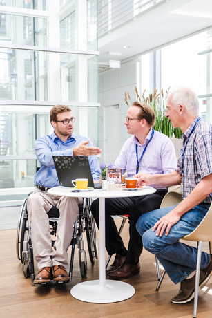 Drei Personen sitzen an einem Tisch. Eine Person sitzt in einem Rollstuhl und erklärt den anderen beiden etwas. Auf dem Tisch befinden sich Getränke und ein offener Laptop.