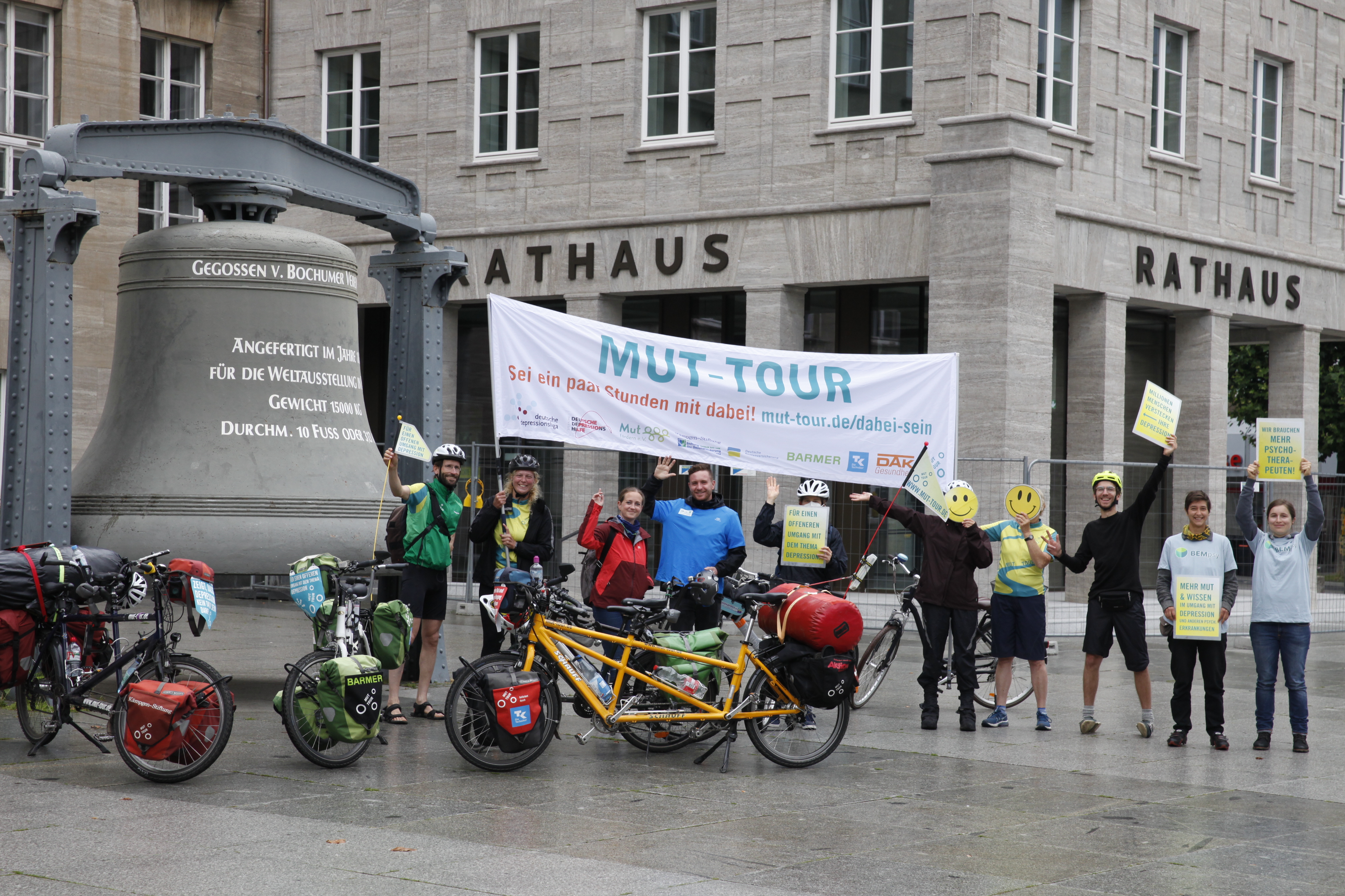 Mehrere Menschen stehen mit Fahrrädern vor einem Rathaus. Sie halten ein Banner und Schilder. Zwei Personen verdecken ihr Gesicht mit Smileys.