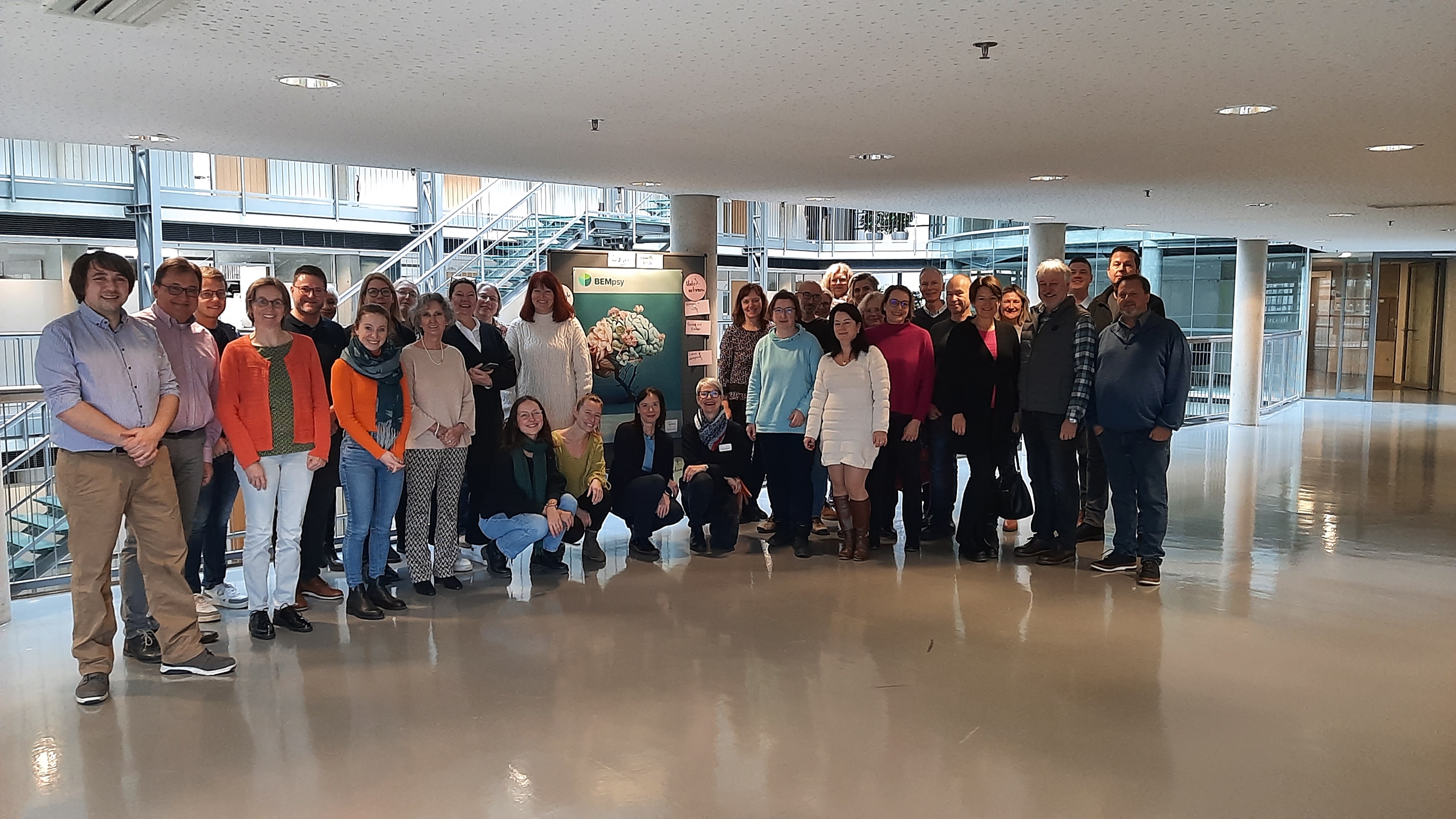 Es ist eine Gruppe von Menschen zu sehen, die in einem Gebäude vor einem Plakat zu BEMpsy stehen.