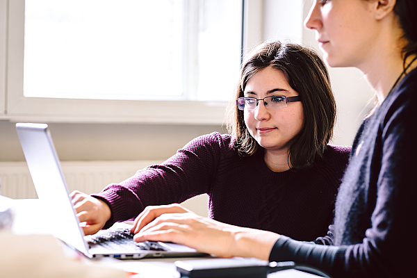 Zwei Personen sitzen an einem Tisch. Eine Person tippt, die andere Person zeigt auf den Laptopbildschirm.