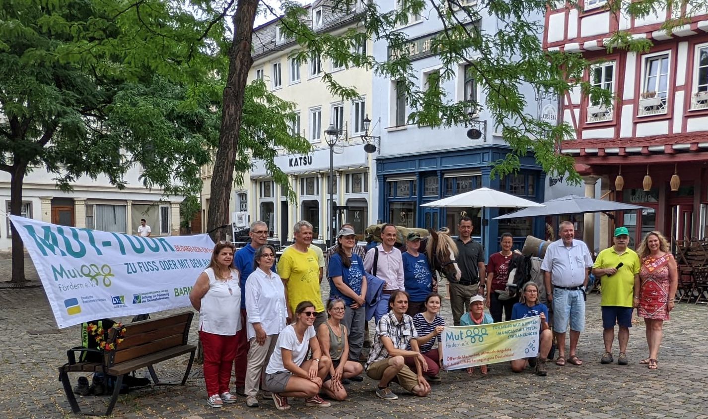 Viele Menschen stehen mit zwei bepackten Pferden auf einem Platz. Sie halten ein Banner hoch, auf dem MUT-Tour steht.