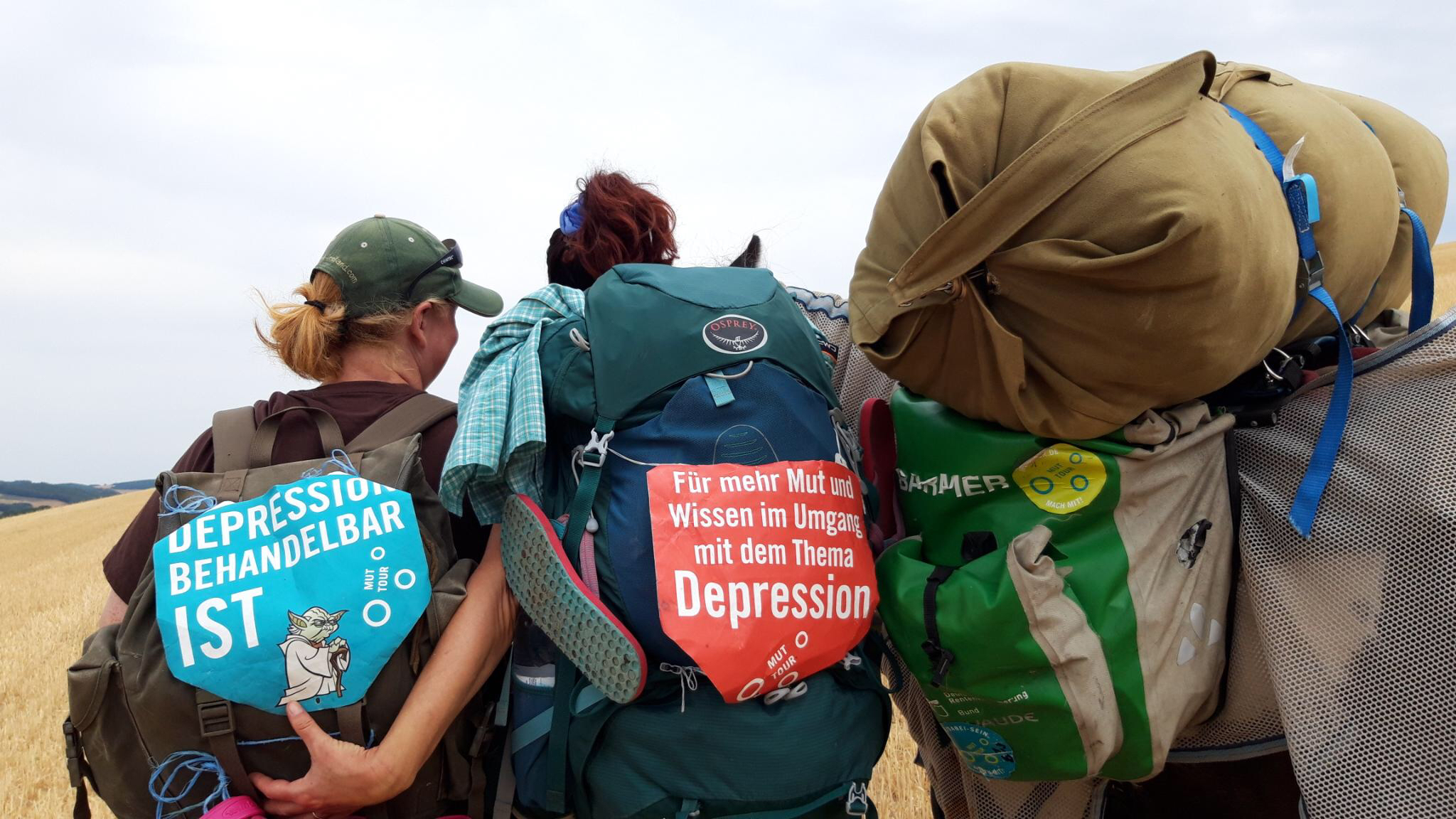 Drei Personen, die von hinten fotografiert wurden und Rucksäcke und anderes Gepäck tragen. An den Rucksäcken hängen Botschaften wie "Für mehr Mut und Wissen im Umgang mit dem Thema Depression".