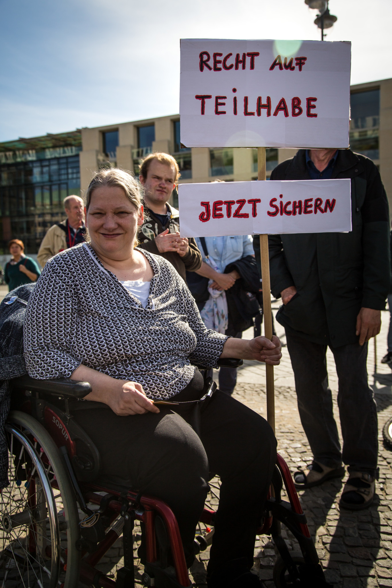 Eine Person sitzt in einem Rollstuhl und hält ein Protestschild, auf dem "Recht auf Teilhabe. Jetzt sichern" steht.