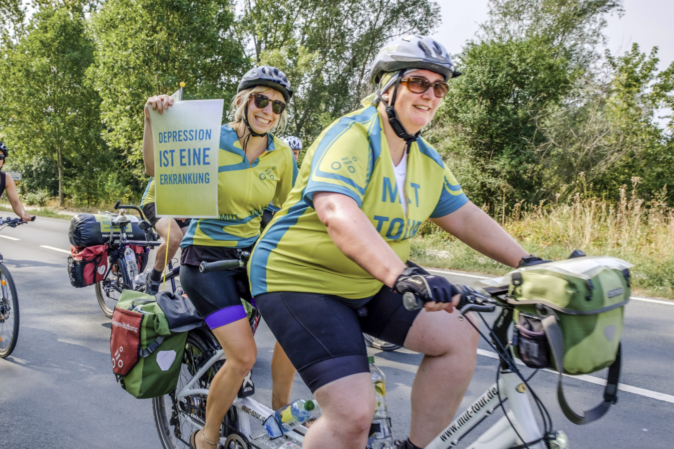 Zwei Personen fahren gemeinsam mit einem Tandem. Die hintere Person hält ein Schild hoch. Darauf steht "Depression ist eine Erkrankung".