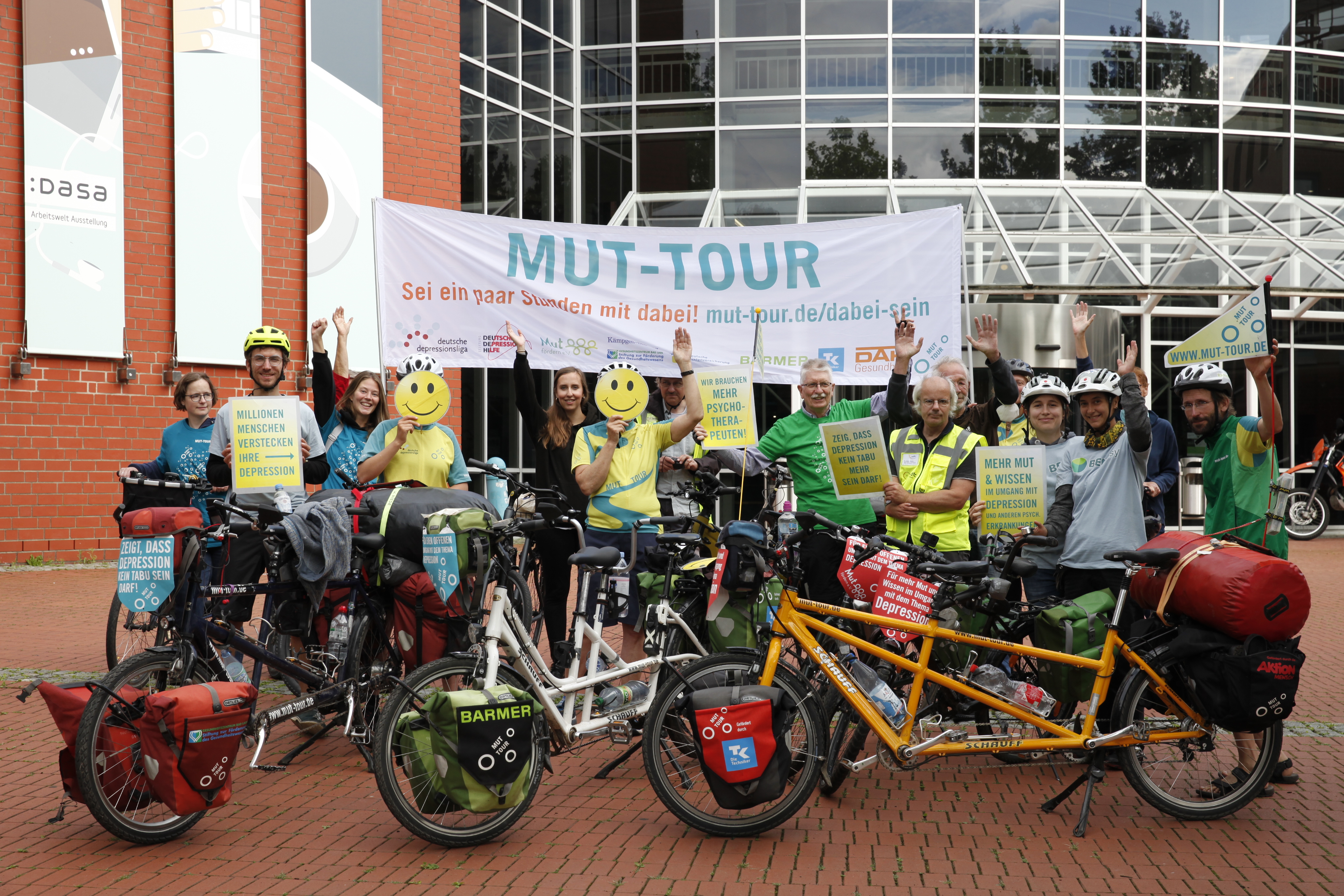 Mehrere Menschen stehen mit Fahrrädern vor der DASA – Arbeitswelt Ausstellung. Sie halten ein Banner und Schilder. Zwei Personen verdecken ihr Gesicht mit Smileys.