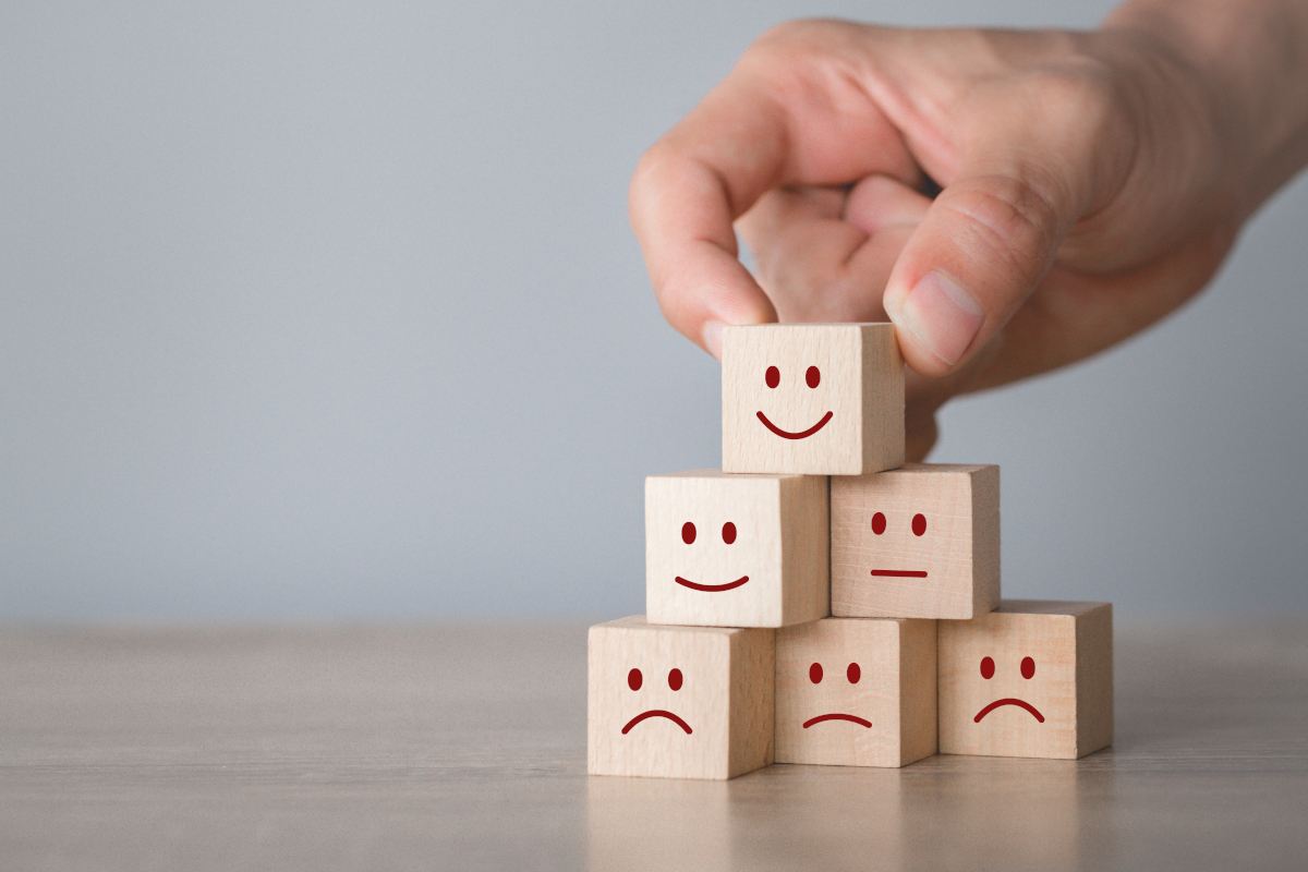 Eine Hand stapelt mehrere kleine Holzklötze zu einer Pyramide. Auf den Holzklötzen sind unterschiedliche Emotionen durch smileys dargestellt. Von glücklich bis traurig.